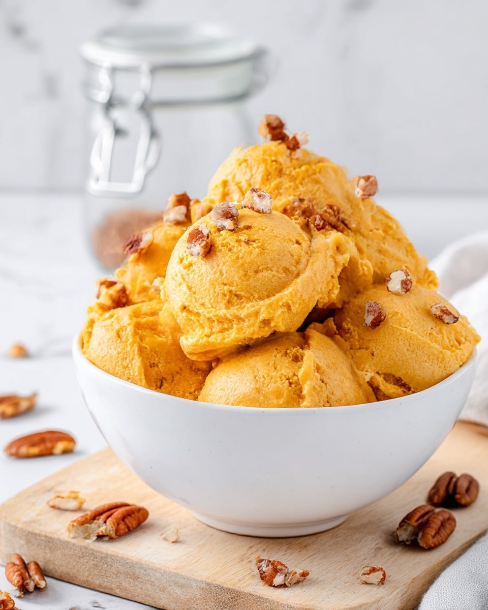 A white bowl is filled with several scoops of smooth, orange pumpkin mousse, each scoop having a soft and creamy texture with a slightly uneven surface. The mousse is topped with small pieces of chopped pecans scattered over and between the scoops. The bowl sits on a light wooden board against a white marbled background, and some pecan pieces are scattered around the base of the bowl. Behind the bowl, there is a clear jar with a silver clasp, slightly blurred. The image shows a close-up view with soft natural lighting. photo taken with an iphone --ar 4:5 --v 7