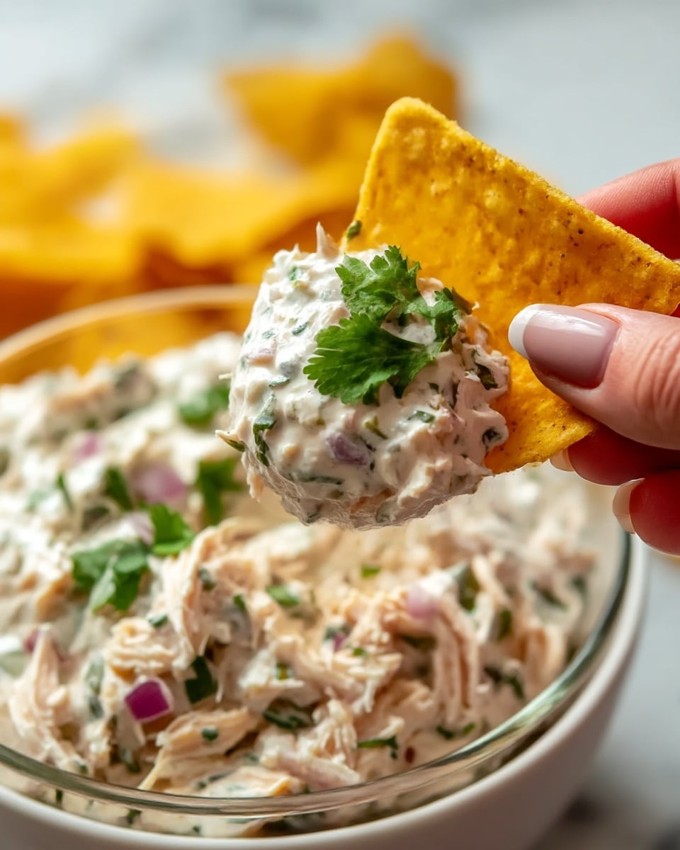 A close-up image shows a yellow corn chip held by a woman's hand with neatly manicured nails. The chip is topped with a thick, creamy white dip mixed with small green herbs and bits of purple onion, with fresh green cilantro leaves added on top. The chip is angled over a bowl filled with the same dip, which looks chunky with visible pieces of shredded chicken and herbs. The bowl is white and sits on a white marbled surface, while blurred yellow corn chips fill the background. photo taken with an iphone --ar 4:5 --v 7