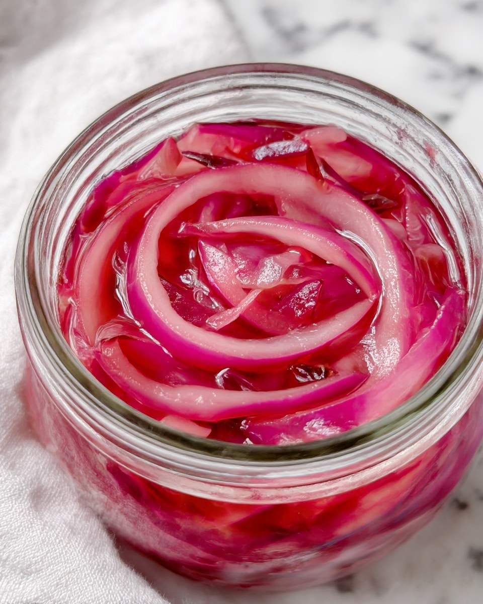 A close-up of a clear glass jar filled with thinly sliced pink and red pickled onions mixed with a glossy, translucent liquid. The onion slices have a smooth, slightly curved texture, layered loosely inside the jar with varying shades of vibrant pink and deep red. The jar is placed on a white marbled surface with a softly blurred white cloth nearby. photo taken with an iphone --ar 4:5 --v 7