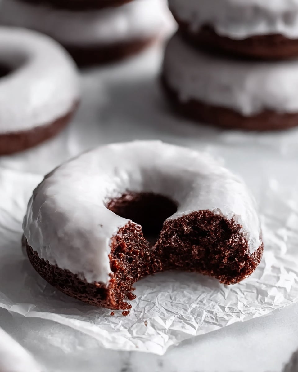 The image shows a close-up of a single chocolate donut with a white glaze coating on top. The donut has a round shape with a hole in the center and looks moist and soft. A bite is taken out of the donut, revealing its dark, rich chocolate inside with a slightly crumbly texture. The donut is placed on crinkled white parchment paper, all sitting on a white marbled textured surface. In the background, there are several more stacked chocolate donuts with the same white glaze, slightly out of focus. photo taken with an iphone --ar 4:5 --v 7