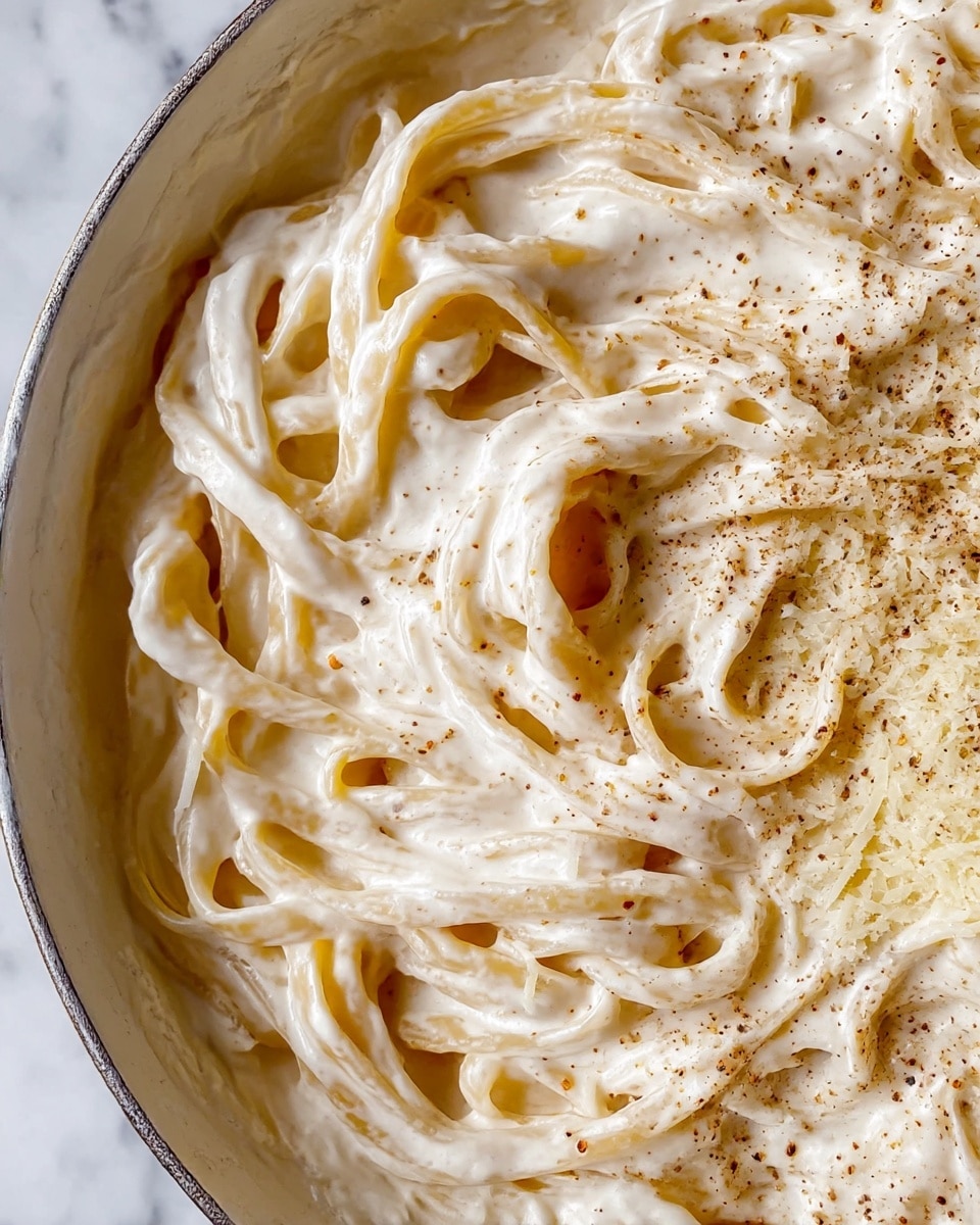 The image shows a close-up of creamy white pasta coated in a thick, smooth white cheese sauce, with a light sprinkle of black pepper evenly spread on the top. The pasta strands are long, flat noodles, twisted and layered on each other, creating a dense but soft texture. Small crumbs of grated cheese are scattered throughout the sauce, adding a bit of grainy detail to the otherwise creamy dish. The pasta is in a white pan, placed on a white marbled surface, enhancing the creamy and rich look of the meal. Photo taken with an iphone --ar 4:5 --v 7