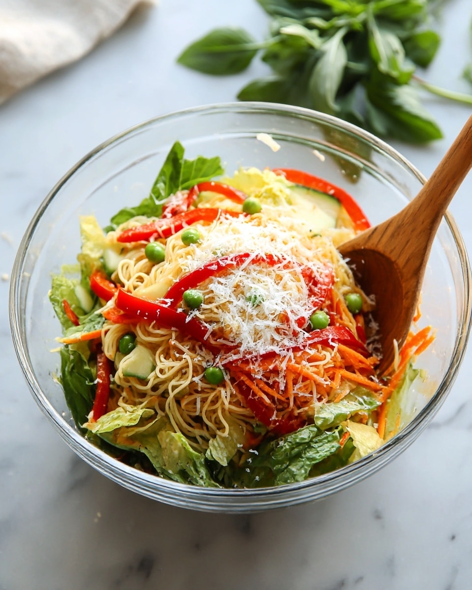 A clear glass bowl filled with a layered noodle salad placed on a white marbled surface. The bottom layer has thin, light yellow noodles mixed with green leafy lettuce. On top of this, there are thin slices of red bell pepper scattered with green peas, sliced cucumber, and shredded orange carrots, adding bright colors. The dish is sprinkled with finely grated white cheese on the top. A wooden spoon is placed inside the bowl resting on the right side. In the background, some green leafy herbs are visible out of focus. photo taken with an iphone --ar 4:5 --v 7
