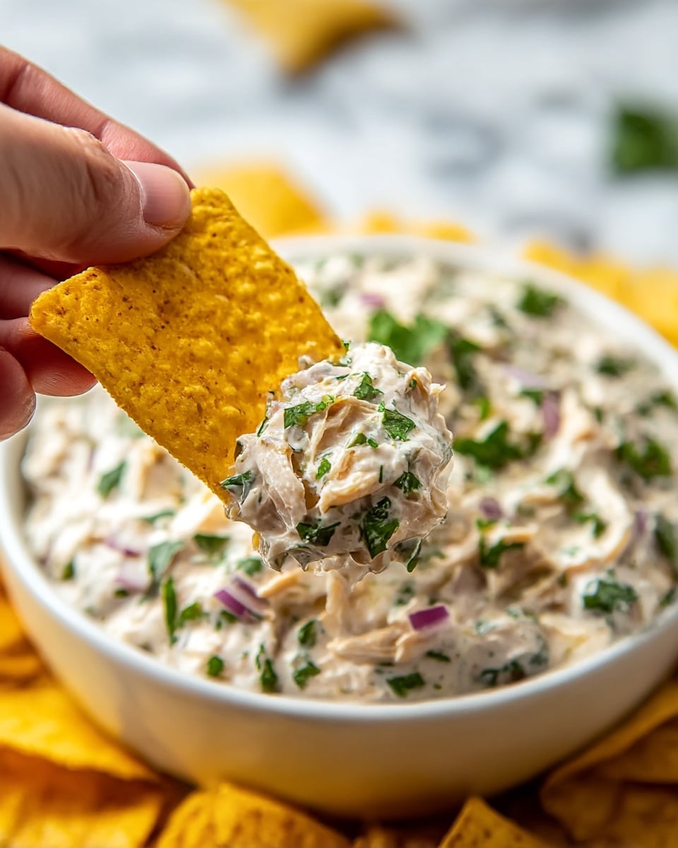A close-up of a white bowl filled with creamy chicken dip, topped with chopped green herbs and bits of purple onion. In the foreground, a woman's hand holds a yellow, textured tortilla chip with a scoop of the creamy dip covering the center. Behind, there are more yellow tortilla chips blurred softly on a white marbled surface. photo taken with an iphone --ar 4:5 --v 7