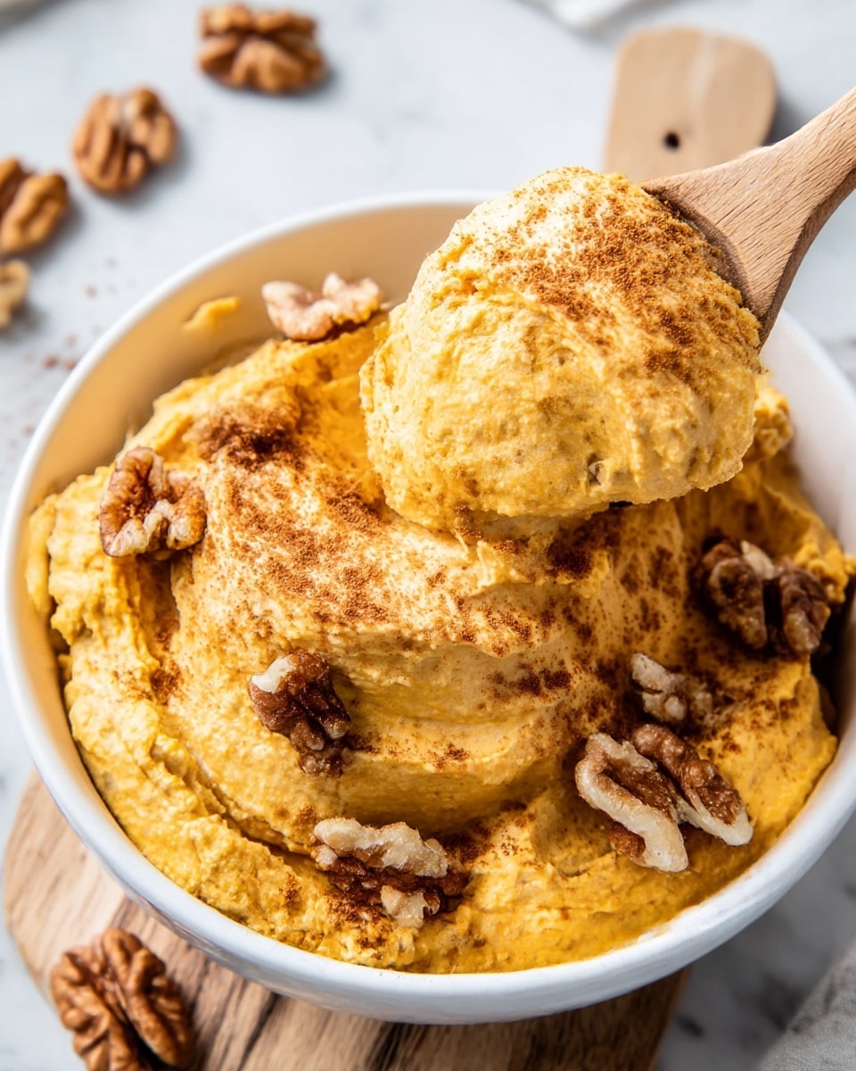 A close-up view of a white bowl filled with thick, creamy orange pumpkin dip layered unevenly with a soft texture. On top and embedded within the dip are scattered halves and pieces of brown walnut halves that add a crunchy contrast. The dip is sprinkled lightly with brown cinnamon powder across the top, adding extra color and texture. A wooden spoon lifts a generous scoop of the dip from the bowl, showing the smooth, slightly chunky consistency. The bowl sits on a white marbled surface with a small wooden board underneath, all softly lit in natural light. Photo taken with an iphone --ar 4:5 --v 7