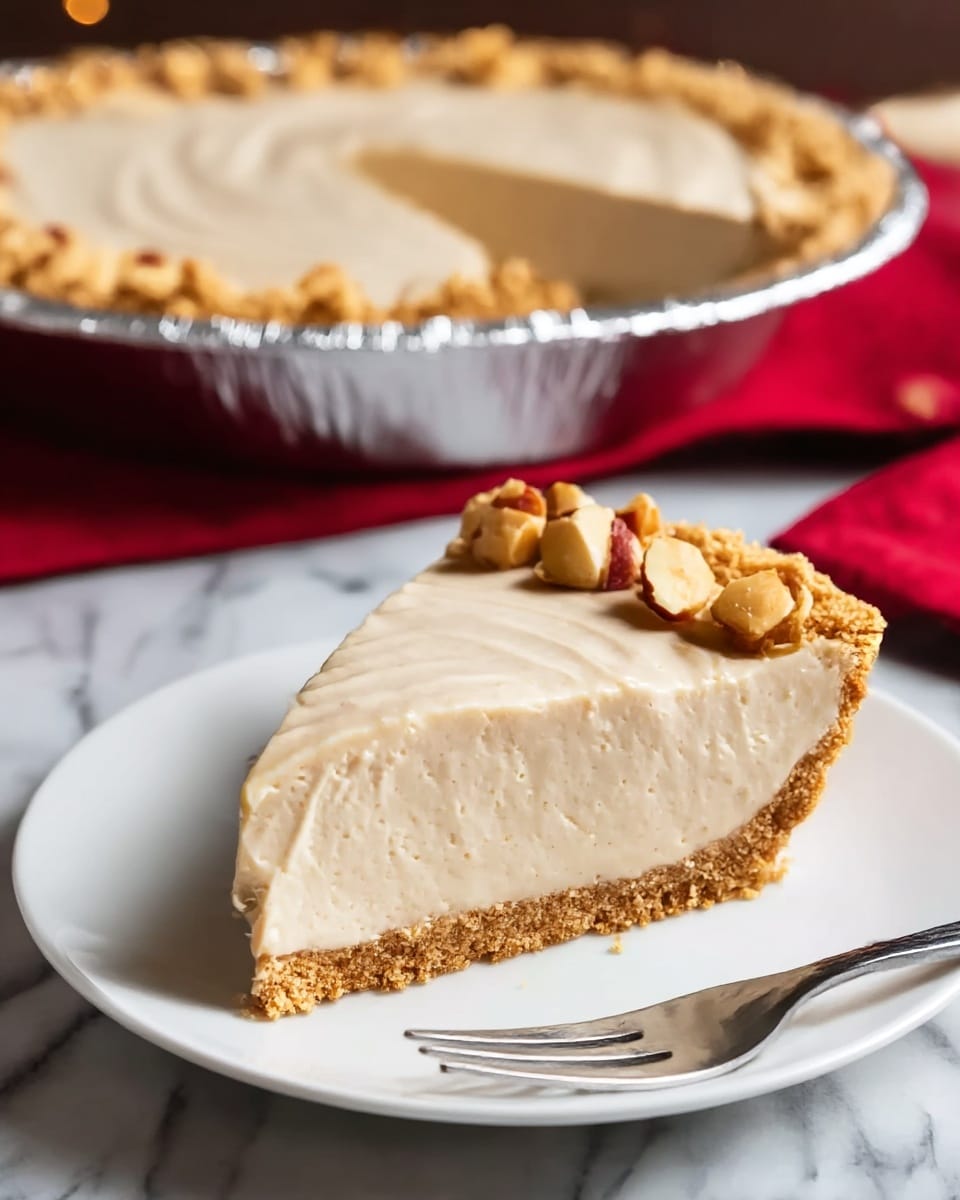 A single slice of light beige creamy pie with a crumbly golden-brown crust at the bottom and edge sits centered on a white plate, topped with a few toasted nut pieces arranged along the upper edge of the slice; the pie filling is smooth with gentle ridges on top, showing a thick, fluffy texture. Behind the plate, the rest of the pie is visible in a shiny silver foil pan with the same crumb crust and creamy filling, placed on a white marbled surface with a red cloth underneath. A metal fork rests beside the slice on the plate. Photo taken with an iphone --ar 4:5 --v 7