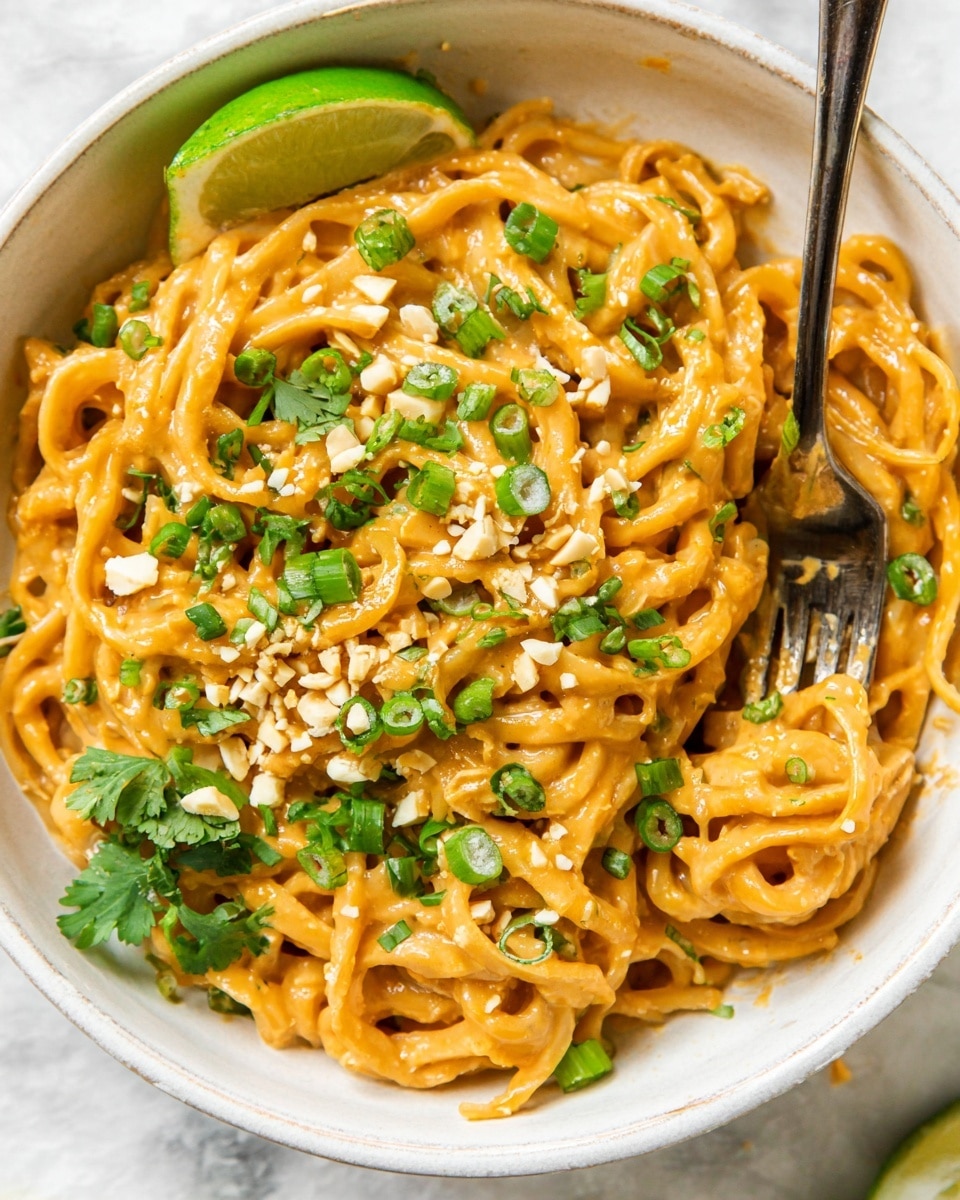 A close-up of a white bowl filled with creamy peanut sauce coated udon noodles. The thick noodles are pale orange and glossy from the sauce, interwoven and piled high. On top, there is a sprinkle of chopped green onions, fresh cilantro, and scattered sesame seeds, adding bursts of green and white. Pieces of crushed peanuts add texture and bits of crunch. A wedge of lime sits on the side, bright green and fresh-looking, next to a silver fork partially buried in the noodles. The bowl rests on a white marbled surface. photo taken with an iphone --ar 4:5 --v 7
