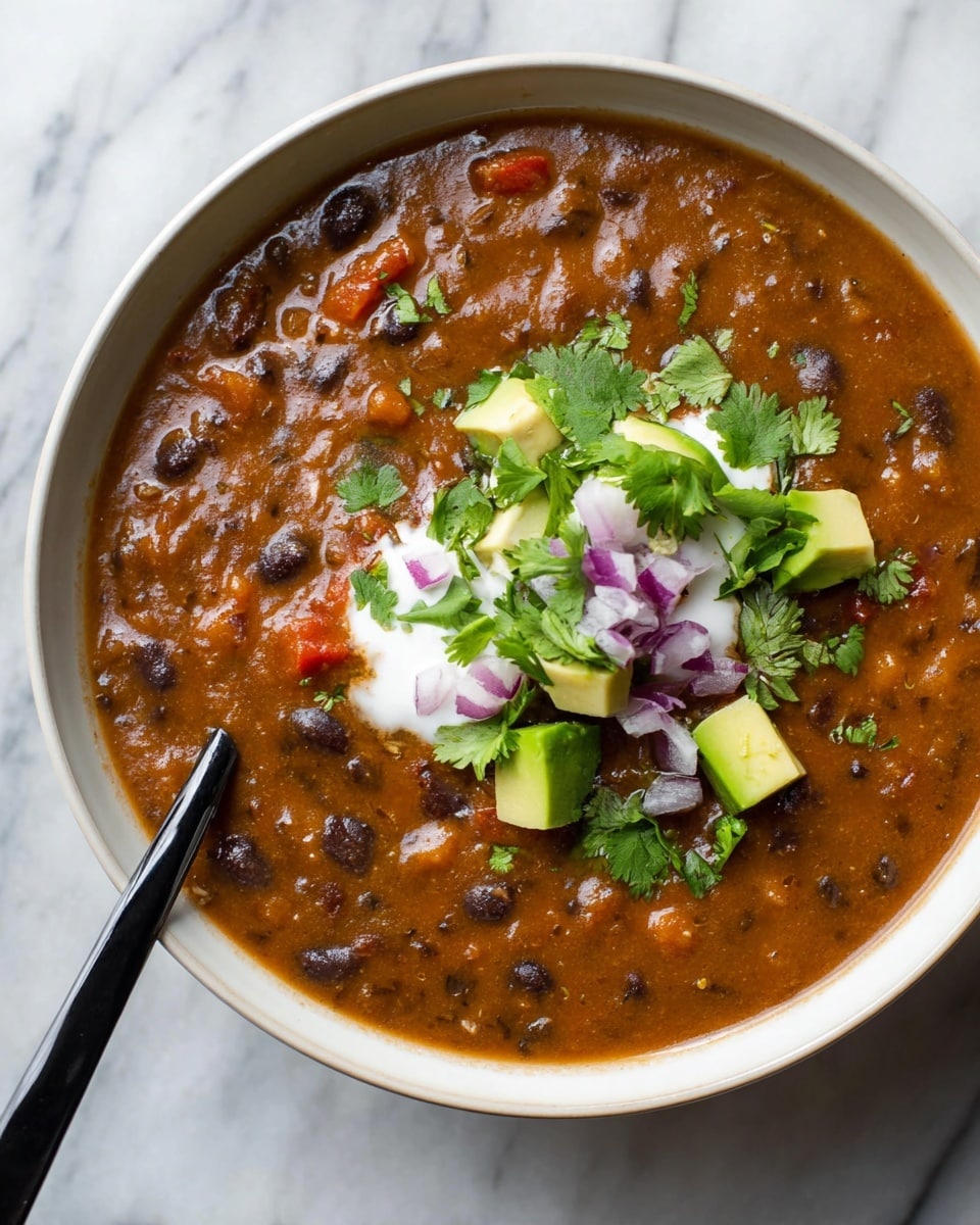 A thick bowl of brown bean soup fills a white bowl, showing a smooth yet chunky texture with visible black beans and small bits of red tomato. On top, there is a dollop of white sour cream, surrounded by green cilantro leaves, small cubes of pale green avocado, and tiny pieces of purple onion. A black spoon dips into the soup on the left side, creating a slight swirl effect. The bowl sits on a white marbled surface. photo taken with an iphone --ar 4:5 --v 7