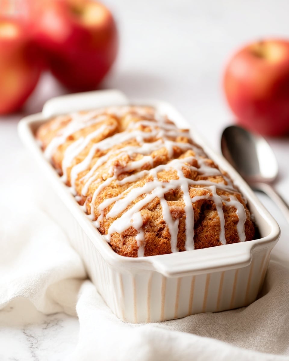 A close-up view of a golden-brown baked bread in a white rectangular ceramic dish, with a crunchy texture on top and white icing drizzled unevenly across the surface. The dish is placed on a soft white cloth, set against a white marbled surface. In the background, there are blurred red apples adding a warm touch, along with a shiny silver spoon resting on the cloth nearby. The lighting is bright and soft, highlighting the bread's texture and icing details. photo taken with an iphone --ar 4:5 --v 7