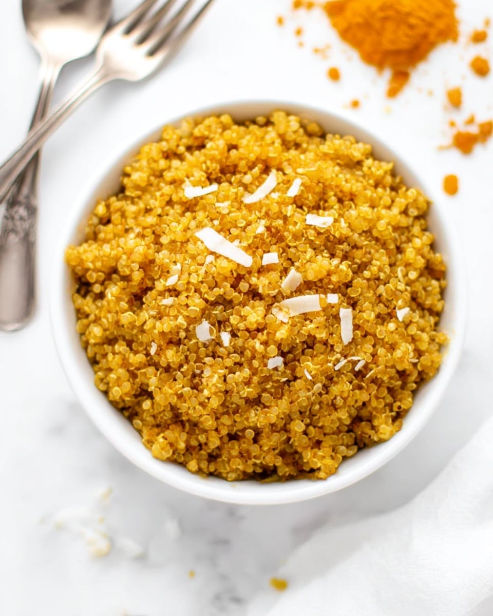 A white bowl filled to the top with cooked quinoa that is yellowish-orange in color, indicating it is seasoned or spiced, sprinkled lightly with small white shreds, possibly cheese or coconut flakes. The texture of the quinoa appears fluffy with each grain separated clearly. The bowl is set on a white marbled surface with a vintage-looking silver spoon and fork crossed nearby, and scattered bits of turmeric powder in the background. photo taken with an iphone --ar 4:5 --v 7