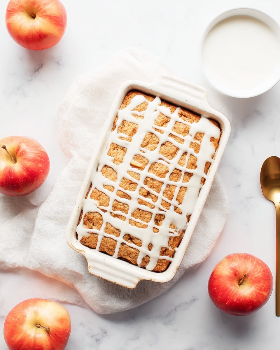 A rectangular baked dessert with a golden brown top layer sits inside a white ceramic baking dish, drizzled with irregular white icing lines across the surface. The dish is placed on a soft white cloth over a white marbled textured surface. Around the dish, there are three red apples with shiny skins and a white bowl filled with white liquid, accompanied by a gold spoon on the right side. photo taken with an iphone --ar 4:5 --v 7