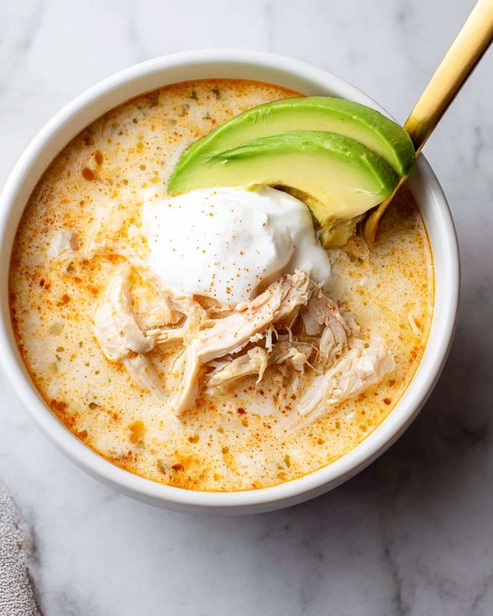 A white bowl filled with creamy chicken soup showing three main layers: the base layer is a light orange creamy broth with small specks of seasoning floating on the surface, the middle layer has shredded pieces of tender light brown chicken partially submerged in the broth, and on top sits a dollop of white sour cream and a slice of bright green avocado placed against the spoon handle, which is metallic gold in color. The bowl is set on a white marbled surface. Photo taken with an iphone --ar 4:5 --v 7