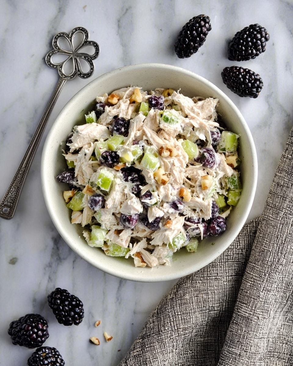 A bowl of creamy chicken salad is shown on a white marbled surface. The salad has three main layers: shredded white chicken pieces mixed in a light, creamy dressing, small bright green celery chunks, and dark purple blackberries scattered evenly throughout. Light brown chopped nuts add texture and are mixed in with the other ingredients. The bowl itself is white and simple in design. Nearby on the marble surface lie a few whole blackberries, and to the right, a textured gray cloth is partially visible. A shiny silver spoon with a flower-shaped cutout design sits near the top left corner of the image. Photo taken with an iphone --ar 4:5 --v 7