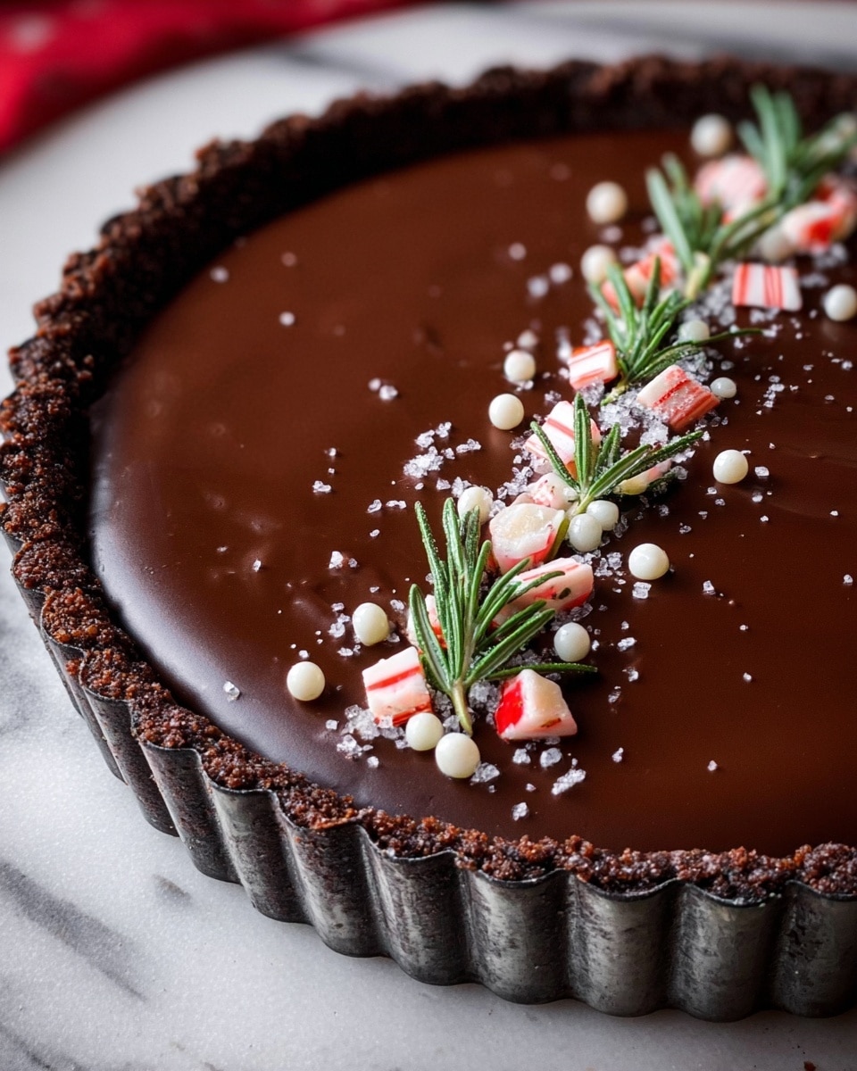This image shows a chocolate tart with a crumbly dark brown crust forming a thick edge around it. The tart has a smooth, glossy dark chocolate layer on top. On the chocolate surface, there is a neat line of small white sugar pearls, pieces of red and white candy cane, and small green rosemary sprigs arranged evenly in the center. The tart is in a metal tart pan with a fluted edge and is placed on a white marbled surface. photo taken with an iphone --ar 4:5 --v 7