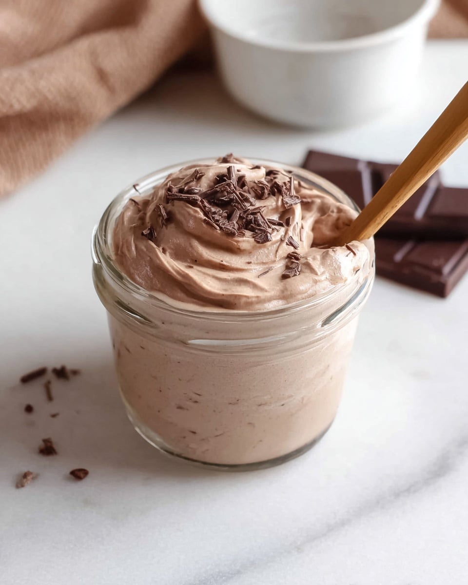 A small clear glass jar filled with creamy light brown chocolate mousse, topped with small chocolate shavings scattered across the surface, with a wooden spoon dipping in from the top right; the mousse looks smooth and thick with soft swirls on top. The jar sits on a white marbled surface with a white bowl and brown cloth blurred in the background and some square pieces of dark chocolate placed nearby on the surface. photo taken with an iphone --ar 4:5 --v 7