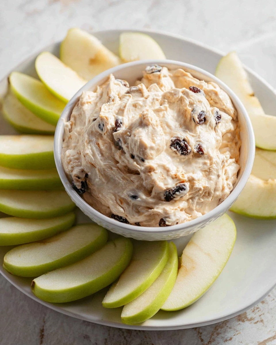 A white bowl filled with a thick, creamy dip that is light beige in color with visible small dark raisins and textured bits mixed in, placed at the center of a white plate. Surrounding the bowl are green apple slices with smooth, shiny skin and pale, juicy interiors arranged loosely around the bowl. The whole setup rests on a white marbled surface. photo taken with an iphone --ar 4:5 --v 7