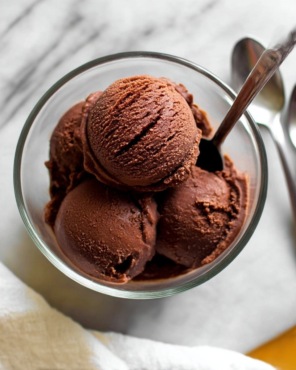 A clear glass bowl holds three scoops of rich dark brown chocolate ice cream, showing a smooth and slightly creamy texture with visible swirls on the top scoop. A silver spoon is placed inside the bowl, resting on the ice cream. The bowl sits on a white marbled surface, adding a light contrast to the dark chocolate color. Another silver spoon is partially visible to the side, and a white cloth is seen at the bottom edge of the image. photo taken with an iphone --ar 4:5 --v 7