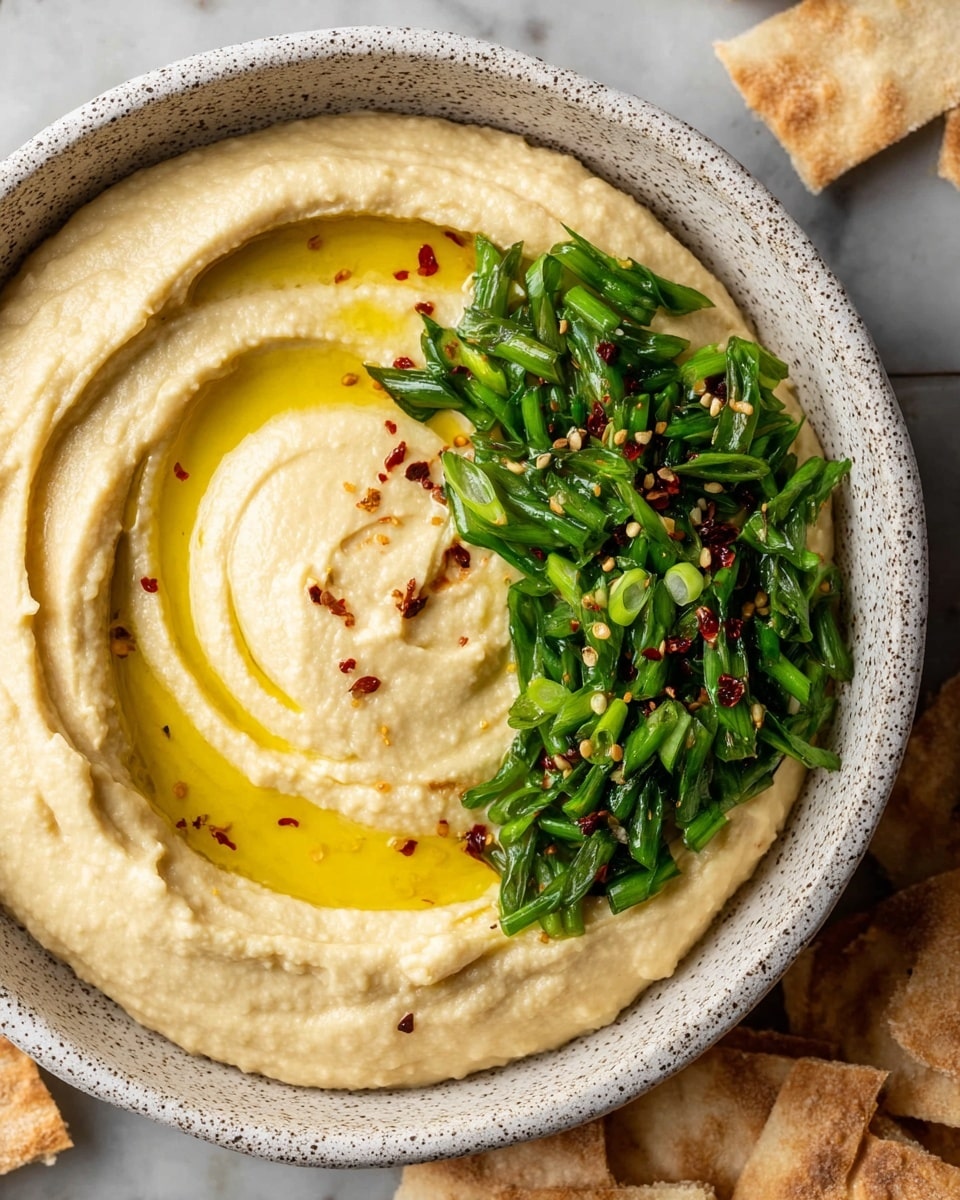 A bowl of creamy beige hummus with a smooth texture forms the base layer. On top, there is a swirl of golden olive oil pooled in the center left, adding shine and richness. To the right of the oil swirl, a layer of chopped bright green scallions spreads in a curved line, providing fresh color contrast. A few tiny red chili flakes are scattered gently across the hummus, adding a touch of red specks for accent. The dish is served in a white speckled bowl placed on a white marbled surface, with some pieces of light brown pita chips resting nearby. Photo taken with an iphone --ar 4:5 --v 7