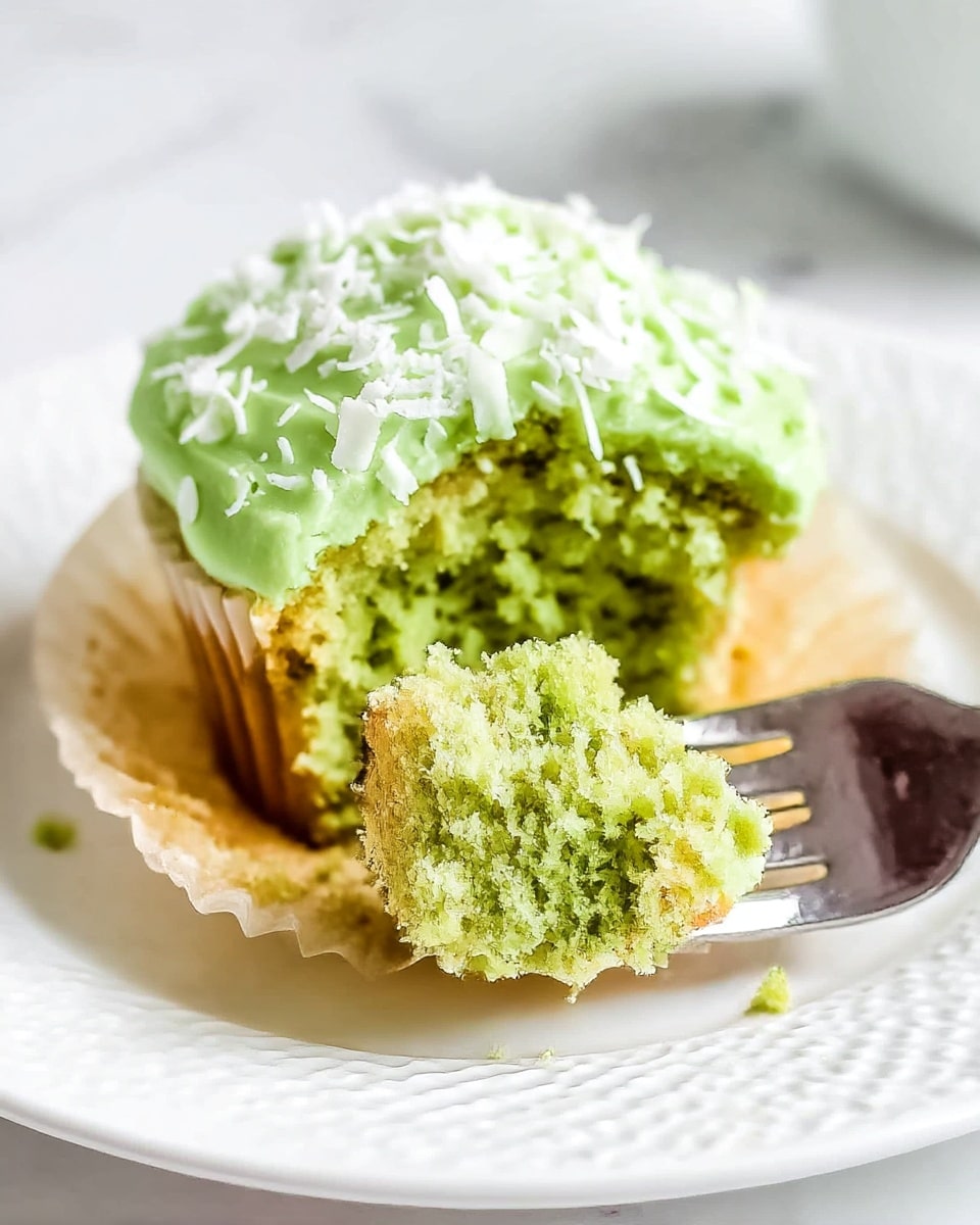 A close-up of a green cupcake with two layers: the top green cake layer is soft and moist, covered with light green frosting decorated with small white coconut flakes scattered unevenly on top; the base layer is a light brown paper cupcake liner. A piece of the cupcake is cut and placed on a white textured plate, held by a silver fork, showing the crumbly texture inside. The background is a white marbled surface, making the bright green color stand out clearly. Photo taken with an iphone --ar 4:5 --v 7