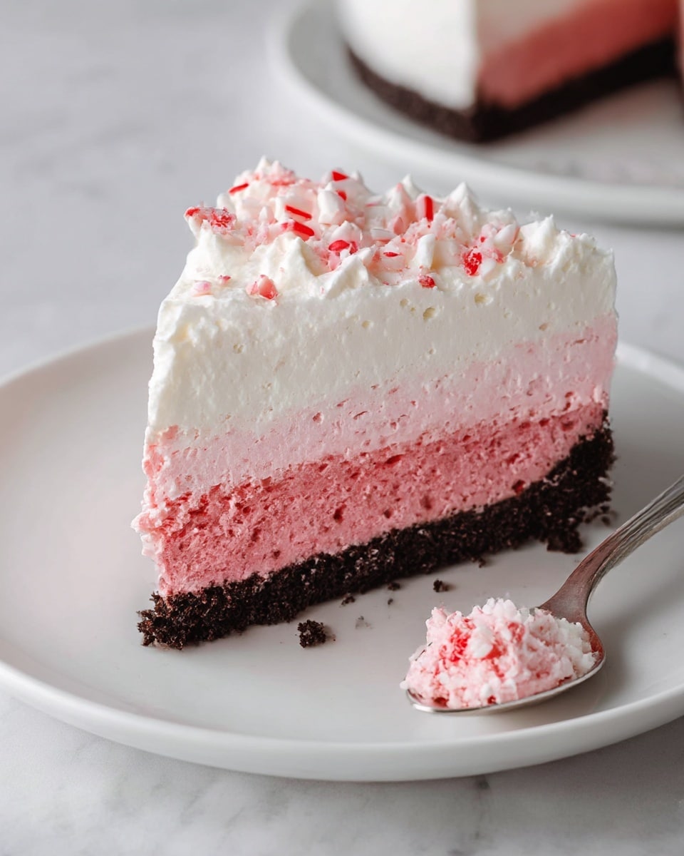 A slice of layered dessert with three distinct layers sits on a white plate over a white marbled surface. The bottom layer is a dark, crumbly crust that looks like crushed cookies. Above that is a thick, smooth pink layer with a mousse-like texture. The top layer is a fluffy white cream, topped with small pieces of crushed red and white candy. A clean bite-sized piece rests on a silver spoon next to the slice, showing the same layers clearly. The lighting highlights the soft textures and colors, creating a fresh and inviting look. Photo taken with an iphone --ar 4:5 --v 7