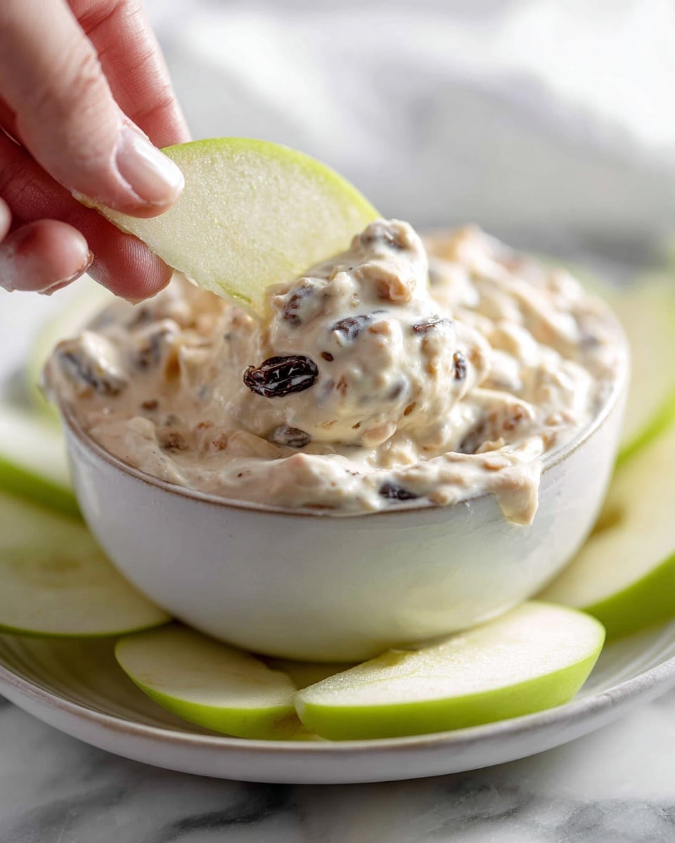 A white bowl filled with a thick creamy dip made of light beige cream mixed with small dark raisins and tiny light brown nut pieces. A green apple slice with smooth, pale green skin and shiny light green flesh is dipped halfway into the creamy mixture. The bowl sits on a matching white plate, and scattered green apple slices are visible on the white marbled surface around it. A woman's hand is holding the green apple slice. The photo taken with an iphone --ar 4:5 --v 7