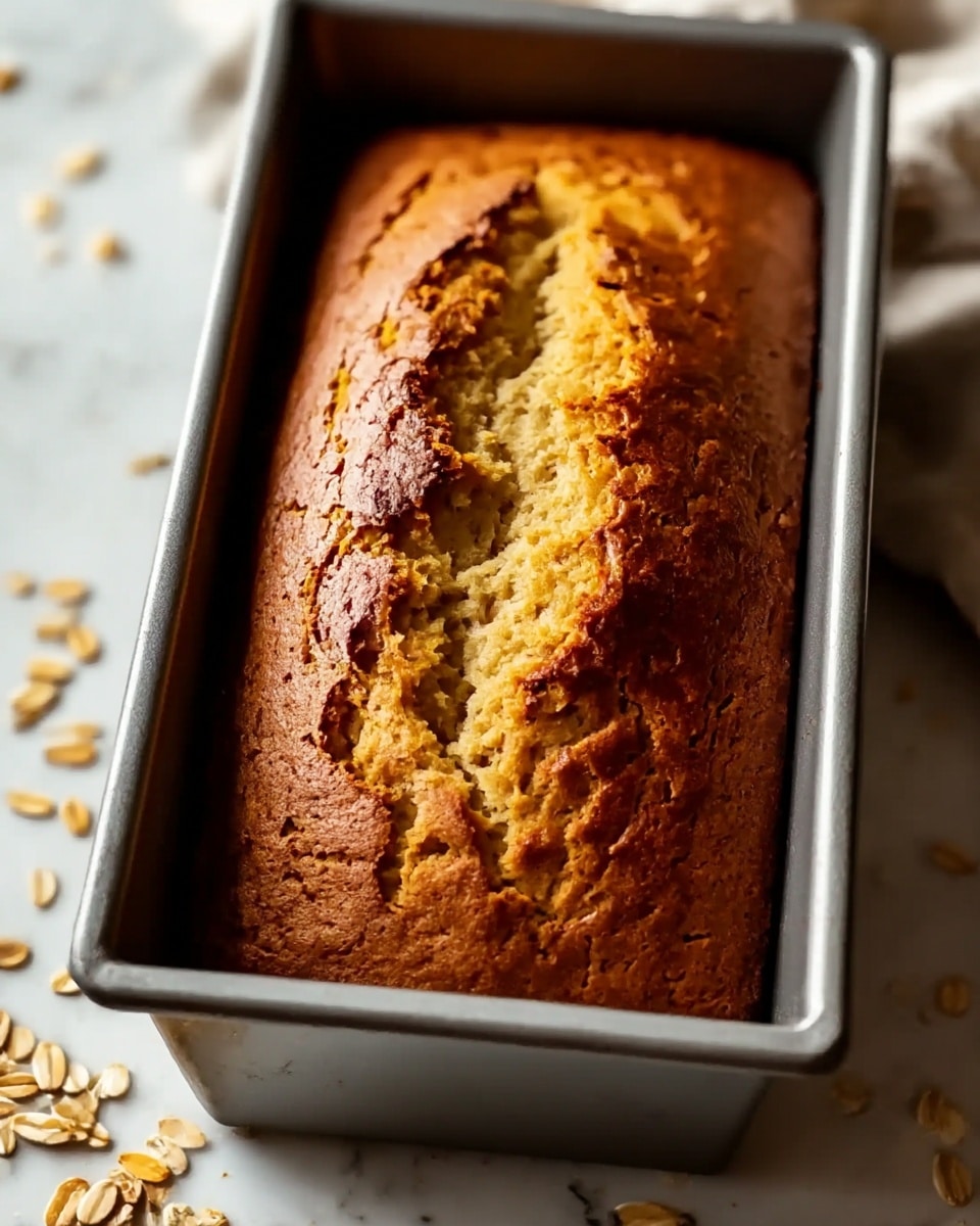 A close-up view of a freshly baked golden brown loaf cake inside a metal rectangular pan, with a cracked top showing a lighter, soft inside. The cake’s surface has a rough, slightly cracked texture with darker brown crust edges. The pan sits on a white marbled surface with a few scattered oats in the corner. Soft natural light highlights the warm tones of the loaf and the cool gray metal of the pan. photo taken with an iphone --ar 4:5 --v 7