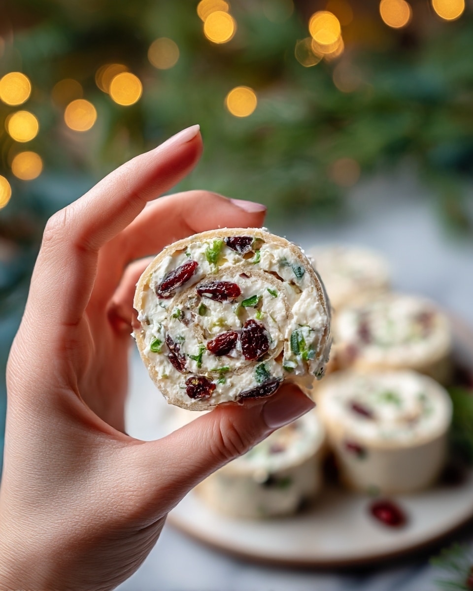 The image shows a white plate holding nine pinwheel sandwiches arranged in close circles, each sandwich consisting of three main visible layers: a light beige soft tortilla base with a slightly rough texture on the outside, a creamy white layer of spread inside, and vibrant red dried cranberries scattered throughout. Bright green chopped chives are sprinkled on top, adding contrast and freshness. The plate's setting includes brown pinecones on the white marbled surface nearby, and out-of-focus warm yellow bokeh lights in the background. Photo taken with an iphone --ar 4:5 --v 7