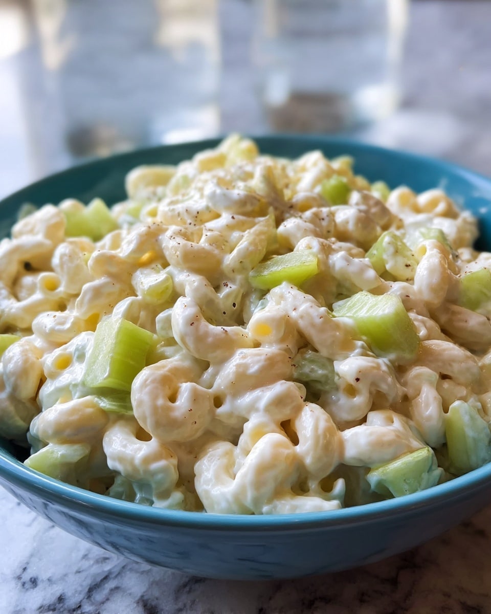 A close-up view of a blue bowl filled with creamy macaroni salad, showing one layer of small curled macaroni noodles coated in a smooth white mayonnaise dressing, mixed with pieces of fresh light green celery cubes scattered evenly throughout. The salad has tiny dark specks of seasoning sprinkled on top, adding contrast against the creamy texture. The bowl is placed on a white marbled surface with a blurred background that includes some glasses of water. Photo taken with an iphone --ar 4:5 --v 7