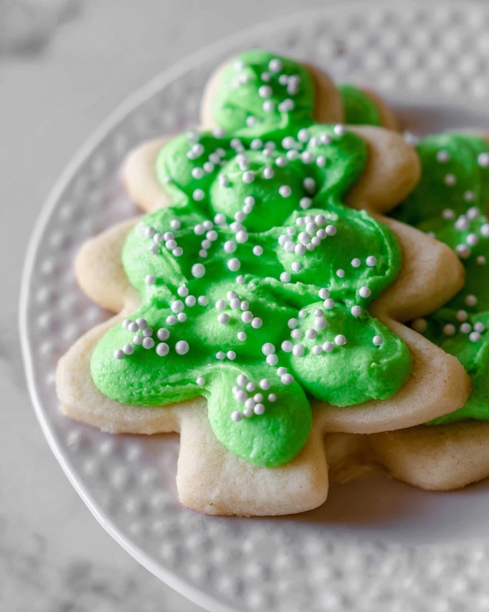 A white Christmas tree-shaped sugar cookie is shown with two layers: the base is a light beige cookie, and the top layer is bright green frosting applied in rounded, uneven dollops to cover the surface. Small white round sprinkles are scattered evenly across the green frosting, giving a festive look. The cookie sits on a white plate with a subtle textured pattern. The background is a white marbled texture. photo taken with an iphone --ar 4:5 --v 7
