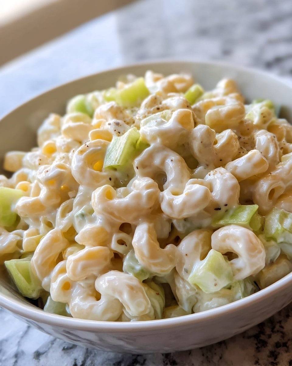 A close-up of a white bowl filled with creamy macaroni salad, showing two main layers: the bottom and middle layers have pale yellow elbow macaroni mixed with small diced light green celery pieces, all coated in a thick, glossy white dressing with a few visible black pepper flakes scattered throughout. The bowl sits on a white marbled surface with a blurred background. photo taken with an iphone --ar 4:5 --v 7