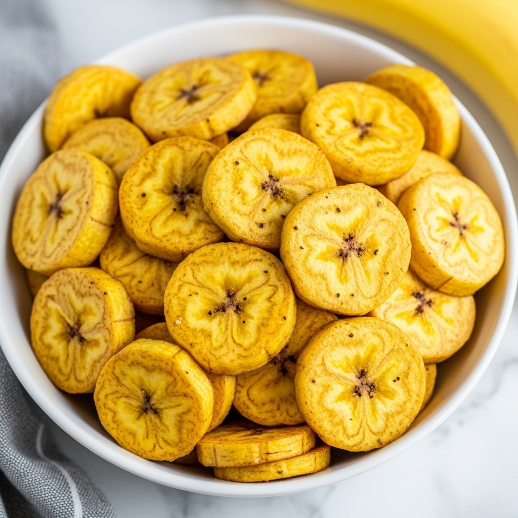 A close-up view of a white bowl filled with many thin, round slices of fried plantain, each slice golden yellow to deep brown with a crisp texture and slight shine. The plantain slices are piled high, showing a mix of smooth, cooked surfaces sprinkled with small dark specks of seasoning. The bowl sits on a white marbled surface with the edge of a yellow banana and a gray and white cloth visible nearby. The photo taken with an iphone --ar 4:5 --v 7