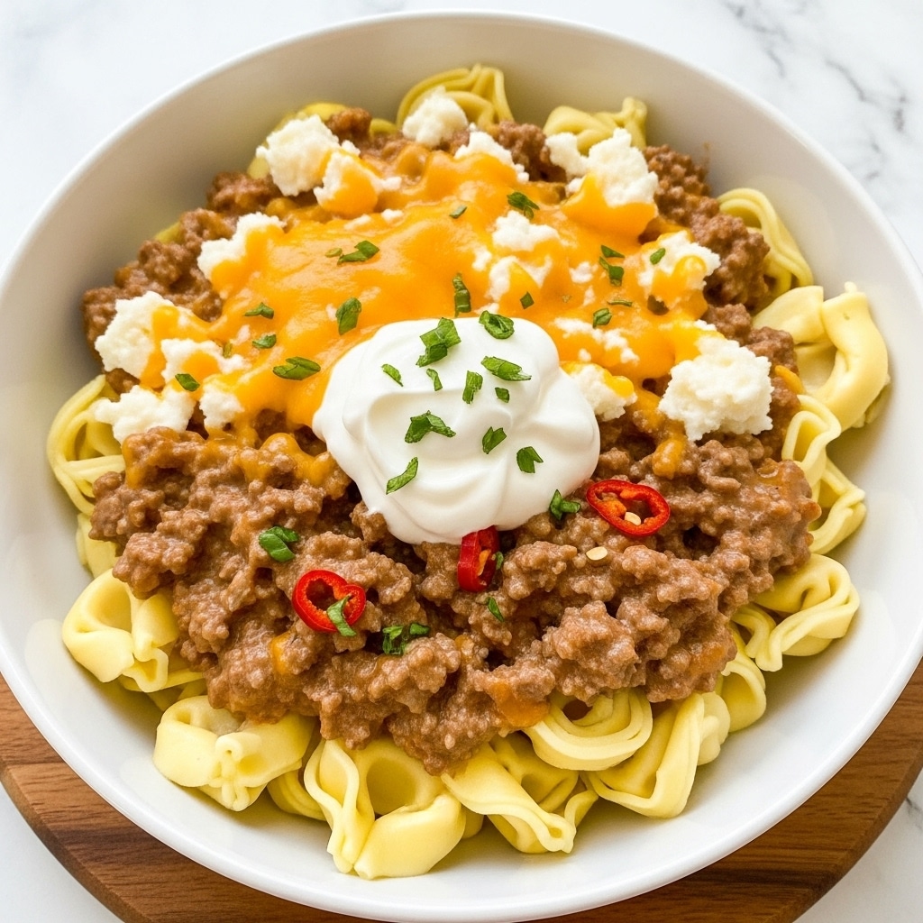 A white bowl filled with creamy cooked tortellini mixed with seasoned ground beef, both coated in a light sauce with visible red pepper flakes. The pasta is a soft yellow with a tender texture, and the beef is a crumbly brown, spread evenly throughout the dish. A generous topping of shredded orange and white cheese covers the tortellini, melting slightly. In the center, there is a dollop of white sour cream sprinkled with small green herb flakes, adding color contrast. The bowl rests on a white marbled surface. photo taken with an iphone --ar 4:5 --v 7