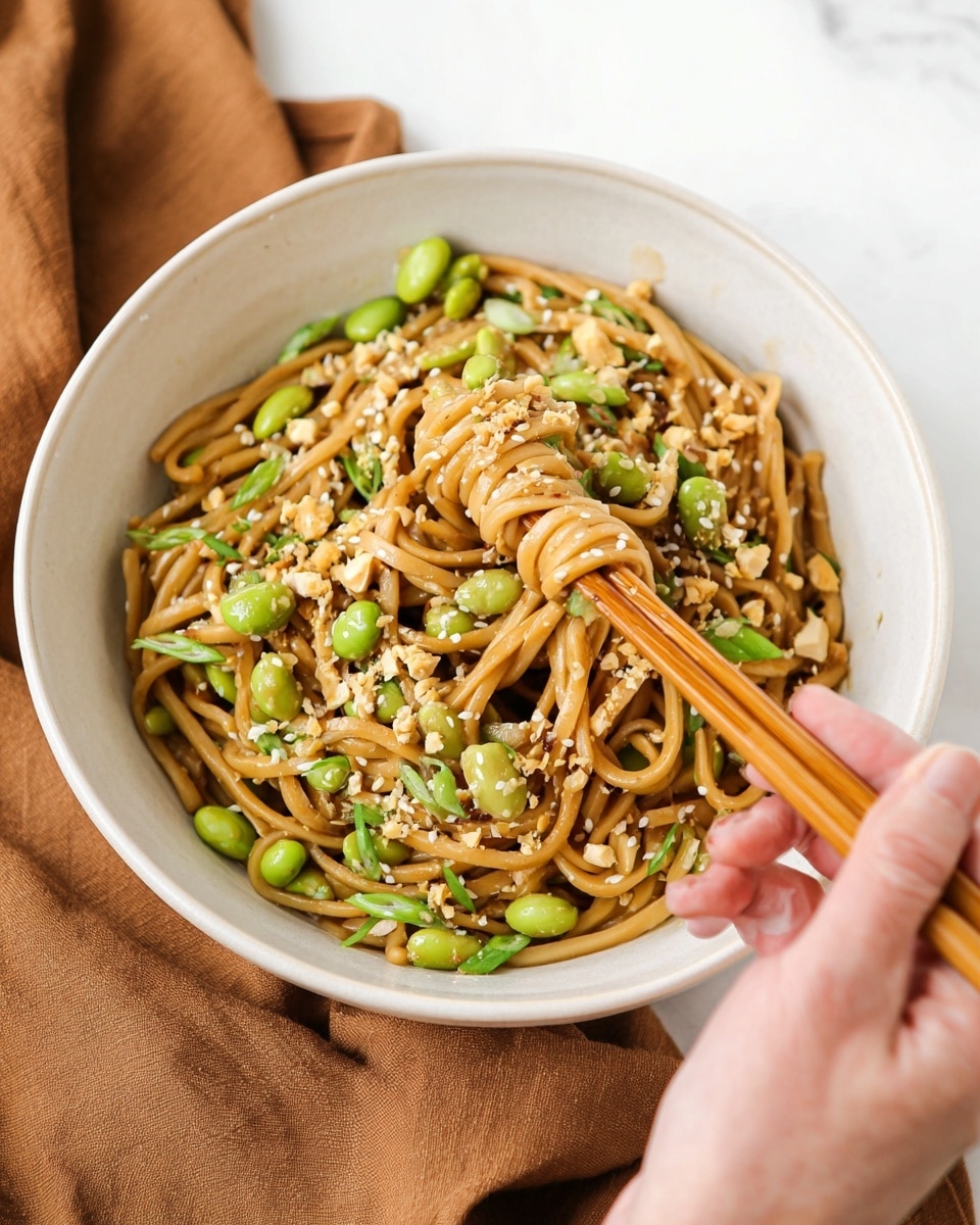 A white bowl filled with thick, light brown noodles mixed with bright green edamame beans and chopped green onions. The noodles have a glossy texture and are topped with small white sesame seeds and crunchy light brown peanut pieces. A woman's hand holding wooden chopsticks is lifting a small bundle of noodles from the bowl. The bowl is placed on a warm brown cloth over a white marbled surface. Photo taken with an iphone --ar 4:5 --v 7