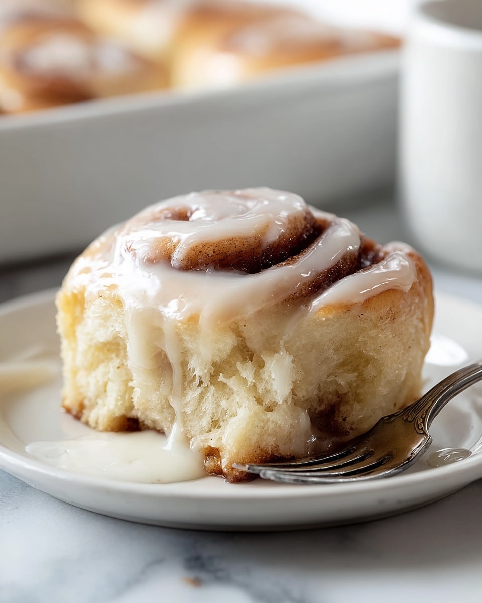 A single cinnamon roll sits on a white plate over a white marbled surface, showing two main layers: a soft, light golden-brown dough spiraled with a darker brown cinnamon filling. The top of the roll is thickly covered with white glossy icing that drips slightly down the sides into a small pool on the plate. A silver fork rests partially under the roll on the right side. In the blurred background, there is a white baking dish holding more cinnamon rolls and a white cup. Photo taken with an iphone --ar 4:5 --v 7