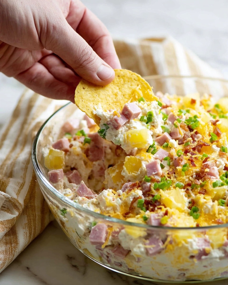 A clear glass bowl holds a creamy, mixed dip made of small pink ham cubes, shredded yellow cheese, white sauce, bright green chopped scallions, and crumbled bacon, layered with bits of light yellow pineapple. A woman's hand is lifting a round yellow corn chip loaded with the dip, showing the mixture's chunky texture and vibrant colors. The bowl sits on a white marbled surface with a beige and white striped cloth blurred in the background. photo taken with an iphone --ar 4:5 --v 7