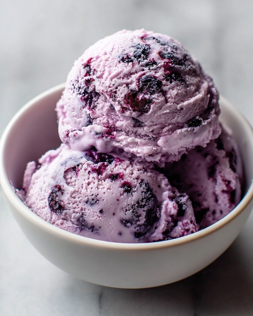 Two scoops of pale purple ice cream with dark purple swirls and chunks fill a white bowl. The ice cream has a creamy and slightly rough texture, showing the berry pieces mixed inside. The bowl sits on a white marbled surface, and the close-up shot captures the rich details of the ice cream’s layers and colors. photo taken with an iphone --ar 4:5 --v 7
