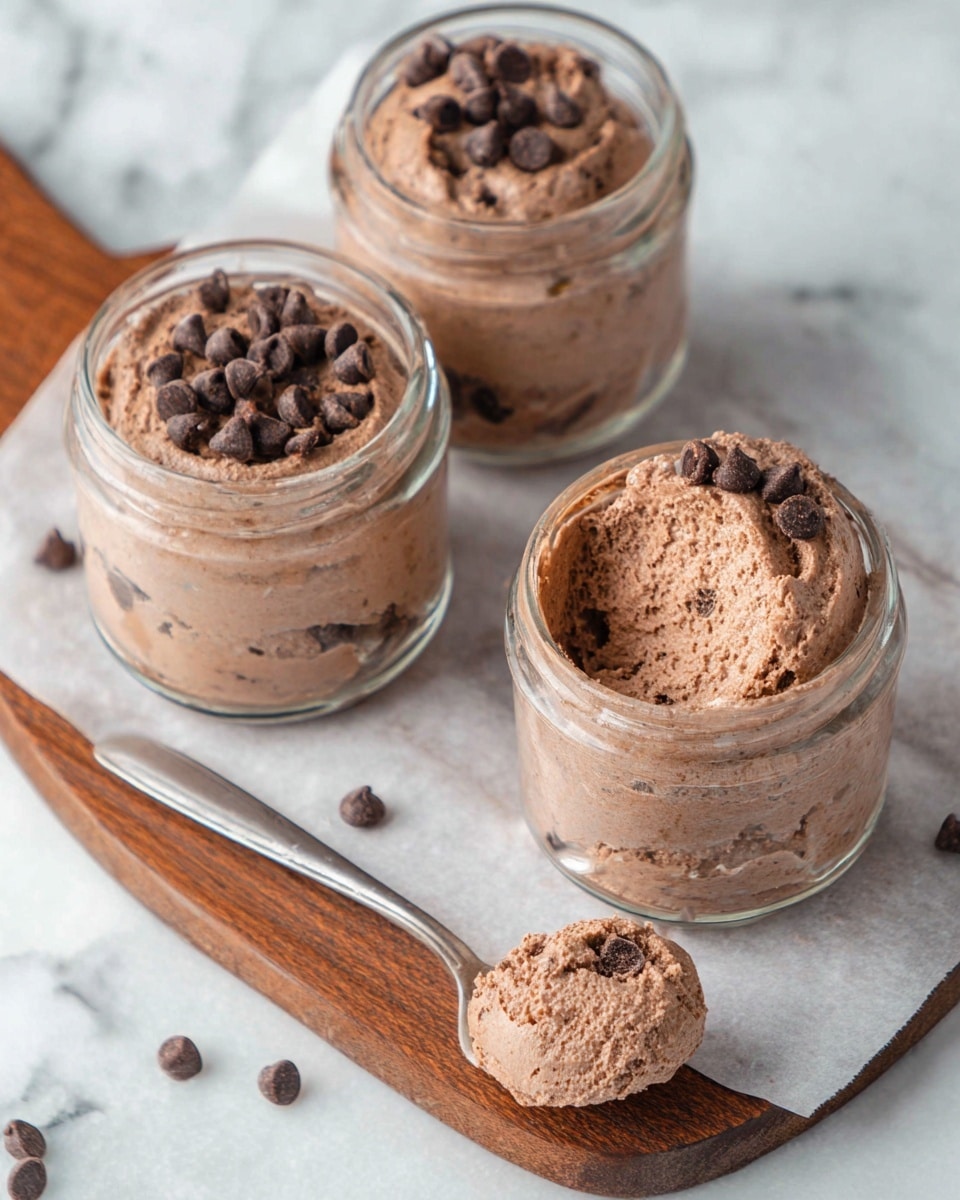Three small glass jars are filled with chocolate mousse that has a thick, fluffy texture and a smooth top layer. The mousse is light brown with visible small dark chocolate chips on the surface and mixed inside. One jar has a partial scoop taken out, showing a creamy inside. Next to the jars, there is a spoon with a large scoop of mousse, also topped with chocolate chips. The jars and spoon rest on a white parchment paper placed on a wooden board, all set on a white marbled surface, and a few chocolate chips are scattered around. Photo taken with an iphone --ar 4:5 --v 7