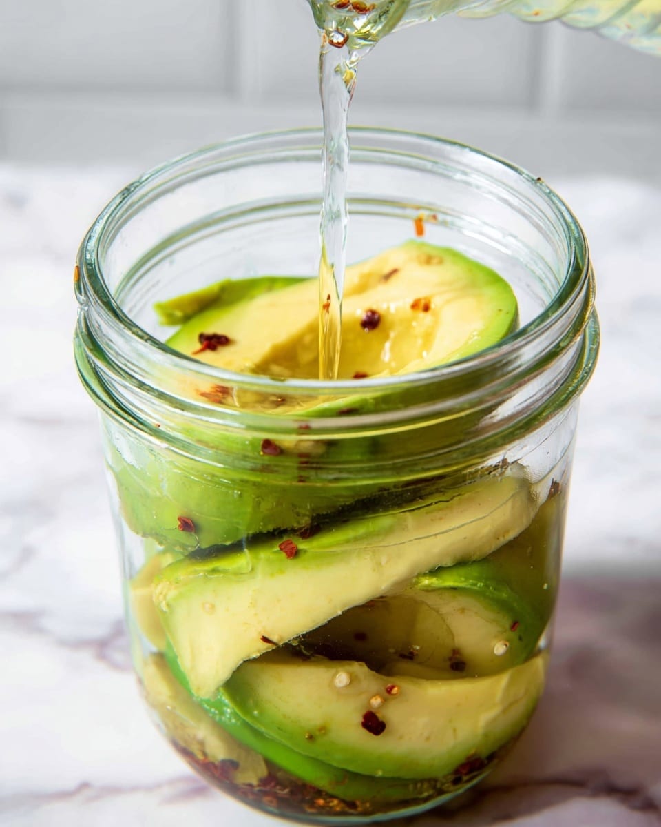 Inside a clear glass jar, several slices of avocado with light green and yellow flesh are stacked, some slices showing darker green edges and slight brown spots. Around the avocado pieces, small bits of red chili flakes float, adding texture and specks of red color. A clear liquid is being poured into the jar from above, visible as a thin stream shining in the light. The jar sits on a surface with a white marbled texture, and the background is softly blurred with a white tone. photo taken with an iphone --ar 4:5 --v 7