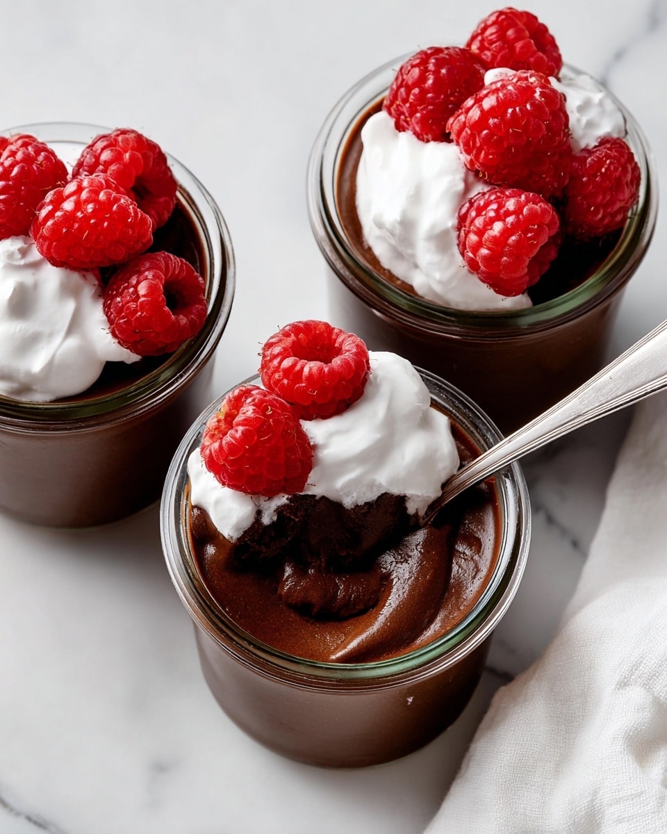 Three small glass jars each contain thick, dark brown chocolate mousse with a shiny, smooth texture filling the jars nearly to the top. Each mousse is topped with a dollop of white whipped cream that contrasts with the dark chocolate underneath. Bright red raspberries sit on top of the whipped cream in pairs or singles, their textured surface and vibrant color adding freshness and visual interest. In the jar at the center right, a silver spoon is partially sunk into the mousse, resting on the edge of the jar, with some whipped cream and mousse mixed around it. The jars are placed on a white marbled surface, with a white cloth visible at the bottom right corner. photo taken with an iphone --ar 4:5 --v 7