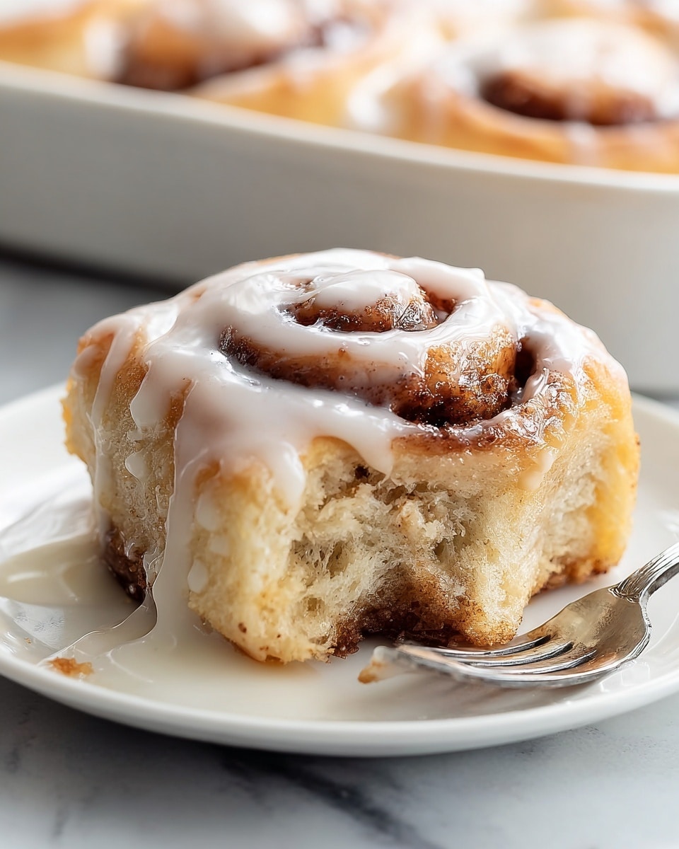 A soft, light brown cinnamon roll sits on a white plate with a glossy layer of thick white icing melting over its spiral top. The roll has a distinct swirl with a darker brown cinnamon sugar filling visible inside, and a moist, fluffy texture that looks freshly baked. A bite has been taken out of the cinnamon roll, showing the inside crumb and cinnamon layer. There are smears of icing pooling around the base of the roll on the plate, and a silver fork rests nearby. The background shows blurred cinnamon rolls in a baking dish, all on a white marbled surface. photo taken with an iphone --ar 4:5 --v 7