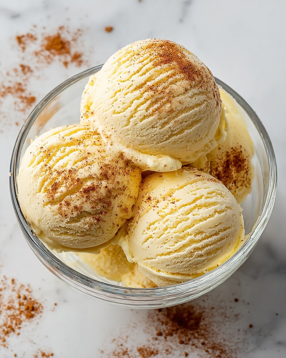 A clear glass bowl holds four scoops of light yellow ice cream with a smooth and creamy texture, each scoop showing curved ridges from the scooping tool. The ice cream is sprinkled with a fine dusting of brown cinnamon powder, particularly on the top and edges of the scoops. The bowl sits on a white marbled surface with scattered specks of cinnamon powder around it. The overall look is clean and simple, focusing on the creamy ice cream and warm sprinkle of spice. photo taken with an iphone --ar 4:5 --v 7