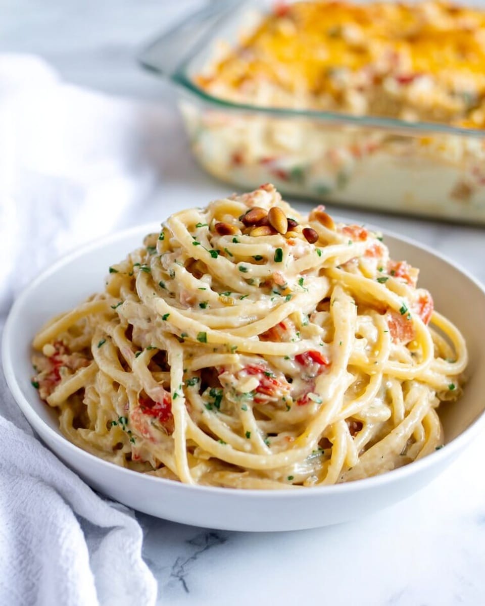 A white bowl filled with creamy spaghetti pasta piled in three loose mounds, showing smooth, pale yellow noodles covered with a rich, light beige sauce speckled with finely chopped green herbs. Small pieces of red tomato and white bits of onion or garlic are scattered throughout the pasta, adding contrast to the creamy layers. A few toasted pine nuts are placed on top, adding texture and a golden color. The bowl sits on a white marbled surface with a white cloth napkin partly visible to the left. In the background, a clear baking dish filled with baked pasta topped with melted golden cheese is out of focus. Photo taken with an iphone --ar 4:5 --v 7