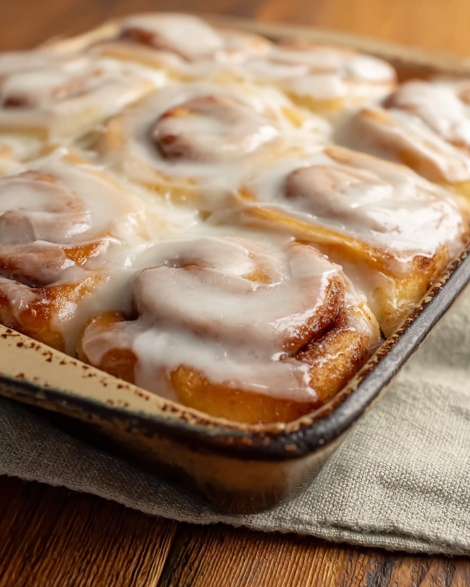 The image shows a close-up of a baking dish filled with freshly baked cinnamon rolls. The rolls are closely packed, with a golden-brown, slightly crispy edge visible along the bottom left corner. Each roll has soft, doughy layers spiraled tightly and covered generously with smooth, white icing that has a slightly glossy texture and drips gently over the edges. The baking dish is rustic and ceramic, and it rests on a neutral beige cloth atop a wooden table with a clear wood grain pattern. The focus is sharp on the front rolls while the background fades softly. Photo taken with an iphone --ar 4:5 --v 7