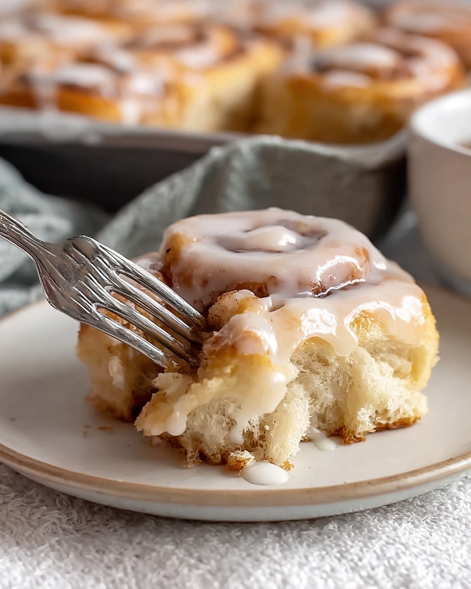 A piece of cinnamon roll with two visible layers sits on a white plate placed on a white marbled textured cloth. The bottom layer is a fluffy, golden-brown dough with a soft texture. The top layer is a thick swirl of cinnamon filling, light brown with darker cinnamon specks, covered by a glossy white icing that looks smooth and slightly melting. A fork is lifting a bite from the cinnamon roll, showing the soft inside and gooey layers. In the blurred background, more cinnamon rolls can be seen in a pan. photo taken with an iphone --ar 4:5 --v 7