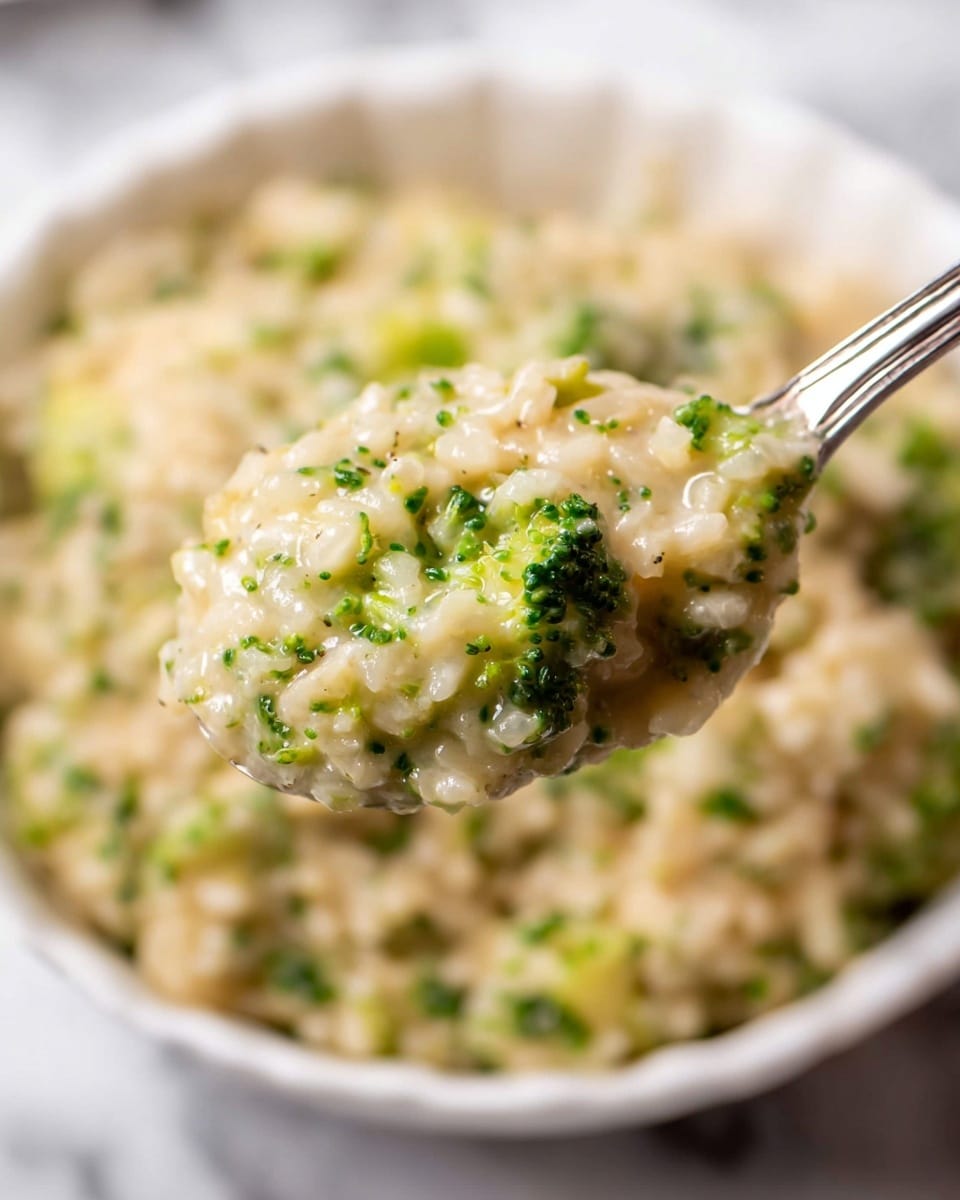 A close-up view of creamy risotto with small green broccoli pieces mixed throughout, showing a soft and slightly chunky texture. The risotto is light beige with tiny bits of bright green broccoli evenly spread inside it. A silver spoon holds a scoop of this mixture, focused clearly at the front, with the rest of the risotto blurred in a white bowl beneath, all set on a white marbled surface. photo taken with an iphone --ar 4:5 --v 7
