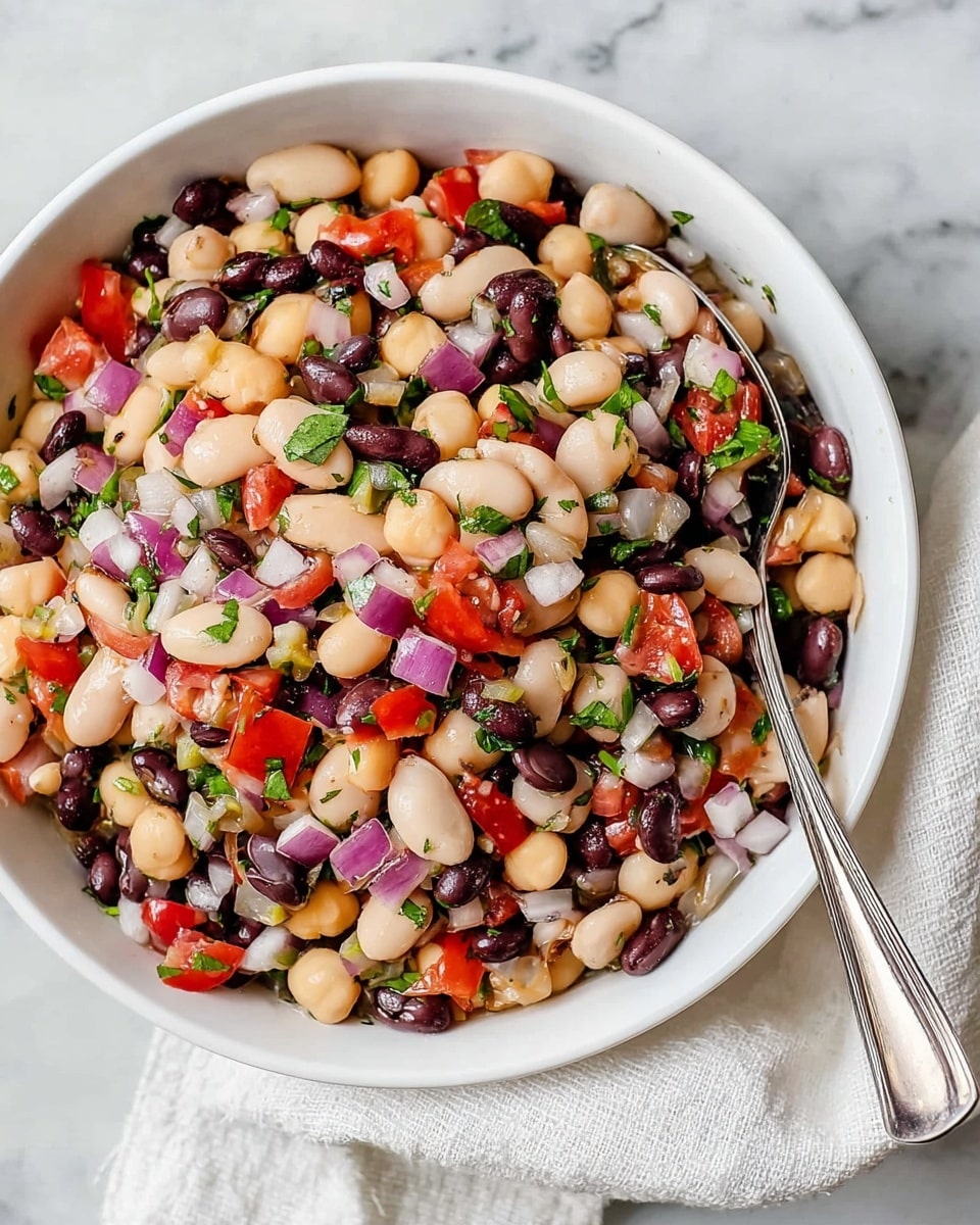 A white bowl filled with a colorful bean salad featuring three main layers: large white beans, small black beans, and round chickpeas, mixed with diced red tomatoes and small cubes of red onion, all scattered evenly throughout. Fresh green herb pieces are sprinkled on top, adding touches of bright color among the beans. A silver spoon rests on the edge of the bowl, partially submerged in the salad. The bowl sits on a white marbled surface with a soft-textured white cloth nearby. photo taken with an iphone --ar 4:5 --v 7
