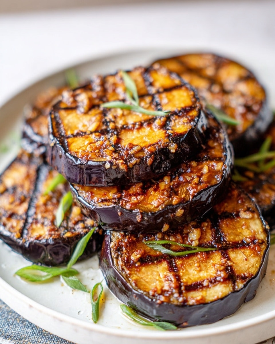On a white plate with a white marbled texture in the background, there are five grilled eggplant rounds stacked together. Each eggplant slice is thick and has a dark purple outer skin. The top of each slice is cut into a crisscross pattern, showing a golden brown charred surface with a slightly crispy texture. The eggplants are coated with a glossy layer of marinade or sauce that has tiny bits of garlic and spices, adding extra texture. Small pieces of green spring onion are scattered underneath the eggplants. The colors are warm with deep browns, golden caramel, and dark purples. photo taken with an iphone --ar 4:5 --v 7