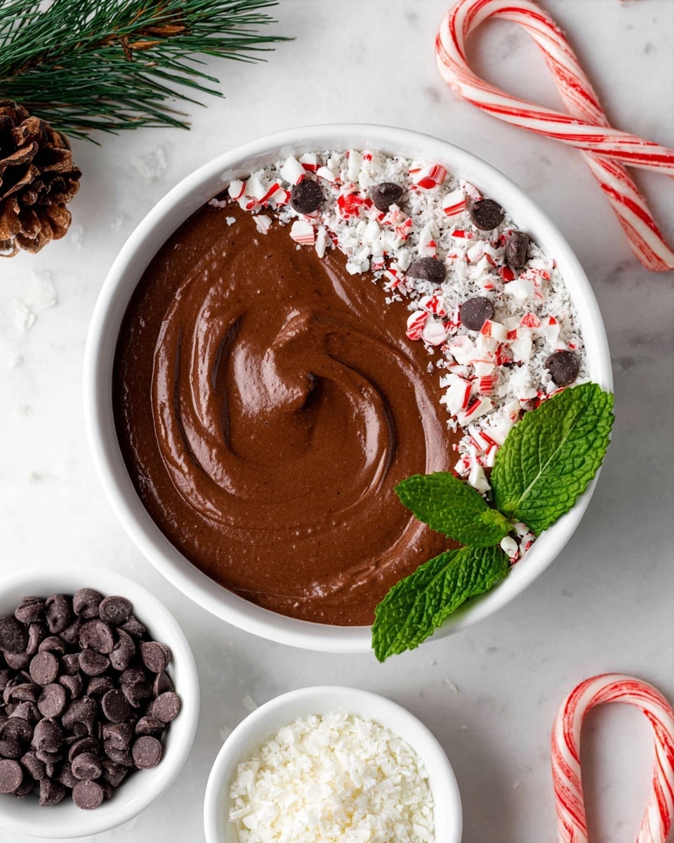A white bowl filled with smooth, dark brown chocolate mousse that has a soft swirl pattern on top. One side of the mousse is topped with a mix of crushed white flakes, small red and white peppermint candy pieces, and dark chocolate chips, creating a textured and colorful crescent shape. A fresh green mint leaf cluster is placed near the mousse surface on the right side for decoration. Around the bowl are small white bowls filled separately with white flakes and dark chocolate chips. The background is a white marbled texture, with a small pine cone and green pine sprigs on the upper left, and several red and white striped candy canes scattered on the right side. Photo taken with an iphone --ar 4:5 --v 7