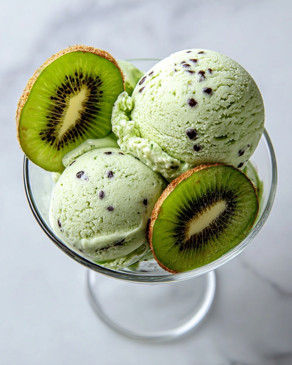 The image shows a glass dish filled with three scoops of pale green kiwi ice cream, each scoop dotted with small black kiwi seeds. Two bright green kiwi slices with dark seeds and brown edges are placed on top and between the scoops, adding fresh color contrast. The glass is clear and sits on a white marbled surface, highlighting the fresh and creamy texture of the ice cream and the juicy look of the kiwi slices. photo taken with an iphone --ar 4:5 --v 7