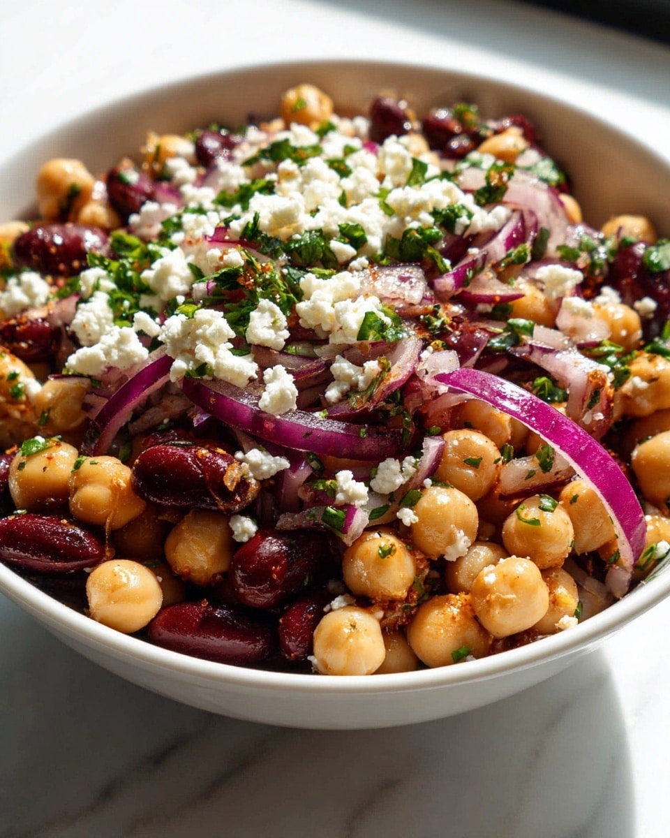 A white bowl filled with a layered chickpea salad sits on a white marbled surface near a bright window. The base layer is made of golden chickpeas and dark black beans mixed together, followed by a layer of finely chopped green herbs scattered evenly. Thin slices of bright purple-red onion rest on top, adding a fresh look. The whole salad is topped with small white crumbles of cheese, giving it a slightly textured appearance. The mix shows a light coating of spices, adding specks of red and brown that blend well with the natural colors of the beans and herbs. photo taken with an iphone --ar 4:5 --v 7