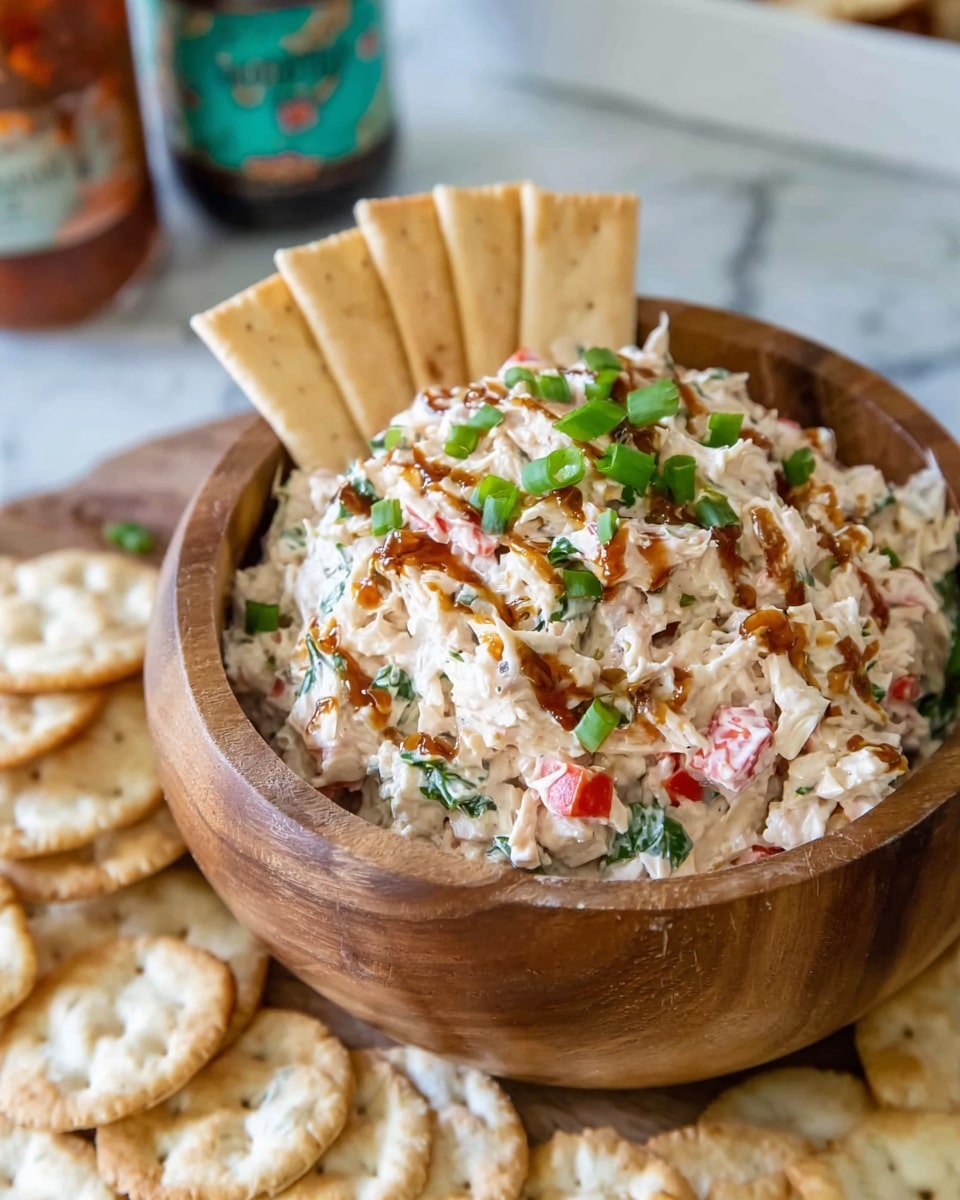 A wooden bowl filled with a creamy chicken salad mixed with small pieces of red bell pepper, green herbs, and onions, topped with a drizzle of brown sauce and chopped green onions. The salad sits in the center with light beige square crackers standing upright behind it inside the bowl. Around the bowl, there are round, slightly bumpy rice crackers spread out on a white marbled surface. In the blurred background, two bottles of sauce and a white rectangular dish are partially visible. Photo taken with an iphone --ar 4:5 --v 7